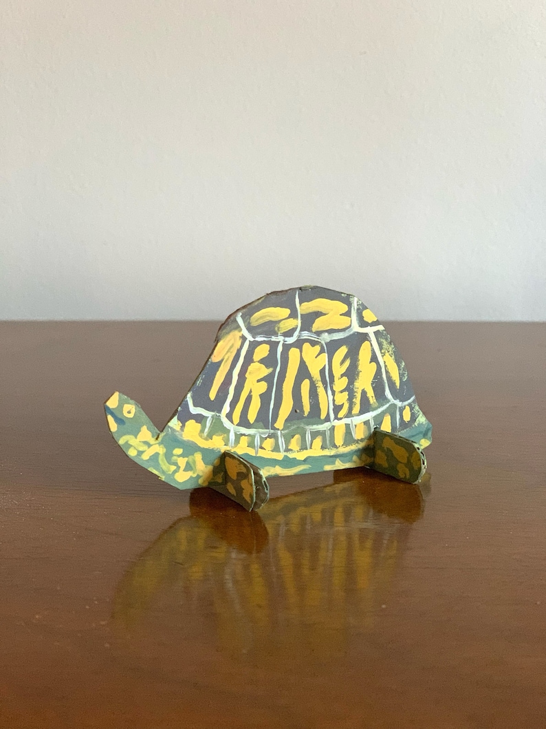 May include: A hand-painted cardboard turtle sculpture with a green, yellow, and black shell. The turtle is standing on a wooden surface.