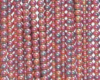 Op de afbeelding: Een streng iriserende, ronde glazen kralen in tinten roze, rood en groen. De kralen hebben een metallic glans en zijn geregen op een doorzichtige draad.
