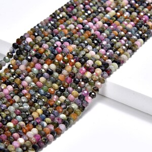 Pode incluir: Um fio de contas de pedras preciosas facetadas multicoloridas. As contas são redondas e têm uma variedade de cores, incluindo verde, rosa, azul e preto. As contas são enfiadas em um fio transparente.