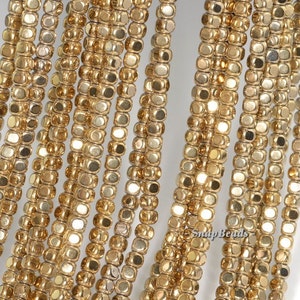 May include: A strand of gold-toned metal beads, each bead is a small square with a faceted surface. The beads are arranged in rows, creating a shimmering effect.