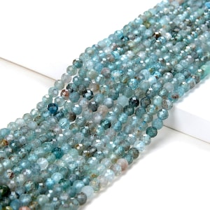 Könnte beinhalten: Eine Strähne aus facettierten blauen Aquamarin-Perlen. Die Perlen sind rund und haben eine hellblaue Farbe mit einigen dunkleren blauen und grünen Einschlüssen. Die Perlen sind auf einem durchsichtigen Faden aufgefädelt.