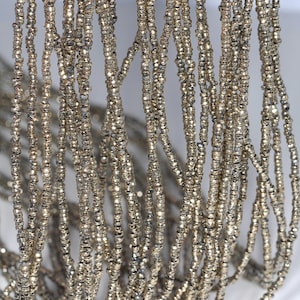 Puede incluir: Una hebra de cuentas de metal plateadas, cada cuenta es facetada y tiene una forma ligeramente irregular. Las cuentas están enhebradas para formar una hebra larga y continua.