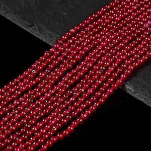 May include: A strand of deep red gemstone beads. The beads are round and smooth, and they are arranged in a neat row. The beads are strung on a clear thread.