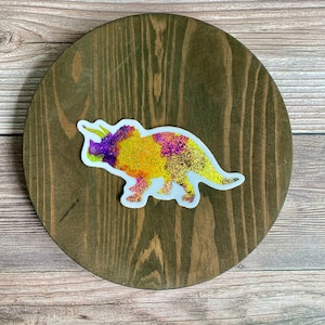 May include: A colorful glitter sticker of a dinosaur, a Triceratops, in shades of yellow, purple, and gold. The sticker is on a brown wooden surface.