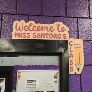 May include: Wooden classroom sign with the words "Welcome to Miss Santoro's" in pink lettering. A pencil-shaped sign with the word "Class" is next to it. The signs are mounted on a purple brick wall.