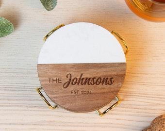 May include: A set of personalized coasters with a two-tone design. The top half is white, and the bottom half is wood-toned, with the text "The Johnsons EST. 2024" engraved. The coasters are in a gold-colored metal holder.