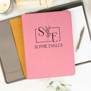 May include: A pink leather folder with a gray flap and a yellow pocket. The folder has a floral design and the initials "S.E." with the name "Sophie Engles" printed below.