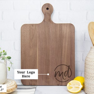 May include: A rectangular walnut cutting board with a handle, measuring 16 inches by 10.5 inches. The board features a circular monogram with the letters "md" and space for a logo. The board is displayed with lemons and wooden spoons.