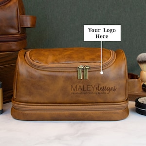 May include: A brown leather travel toiletry bag with a zipper closure and a separate compartment. The bag has the text "MALEY designs Personalized Clothing & Gifts" printed on the front.
