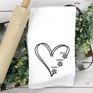 May include: White kitchen towel with a black heart design and paw prints. The names Milo, Cooper, and Jasper are printed inside the heart. A wooden rolling pin and greenery are in the background.