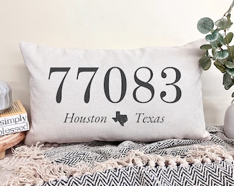 May include: A rectangular beige pillow with the number "77083" printed in dark gray, along with "Houston Texas" and a Texas outline. The pillow is on a patterned blanket.