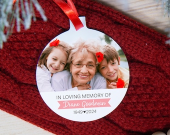 May include: A white round Christmas ornament with a photo of three people, a woman and two children. The ornament has a red ribbon and the text "IN LOVING MEMORY OF Diane Goodman 1949 ♥ 2024".