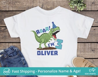 May include: White t-shirt with a green dinosaur wearing a blue party hat. The dinosaur is roaring and holding a blue number 3. The text "Roar! I'm Oliver" is printed on the shirt.