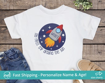 May include: White t-shirt with a blue circle design featuring a rocket ship with a yellow flame and the text "my first trip around the sun". The rocket ship has a window with the name "Wyatt" written on it.