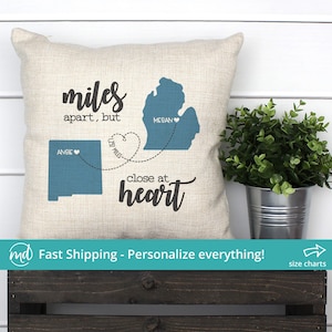May include: A beige throw pillow with a blue outline of Michigan and New Mexico. The pillow has the text "miles apart, but close at heart" with the distance between the states listed. The names "Megan" and "Angie" are written next to the states.