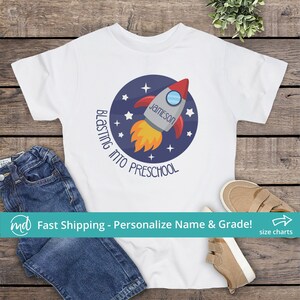 Puede incluir: Camiseta blanca con un gr&aacute;fico circular azul de un cohete con llamas y estrellas. El texto "Blastin' into Preschool" est&aacute; escrito alrededor del c&iacute;rculo. El nombre "Jameson" est&aacute; escrito en el cohete.