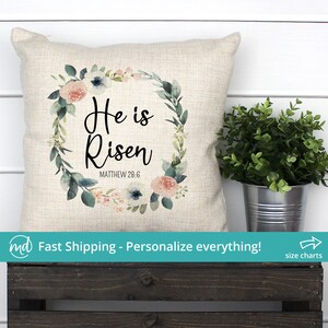 May include: A beige throw pillow with a floral wreath design and the text "He is Risen" with the scripture reference "Matthew 28:6".