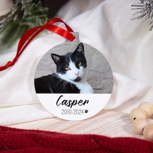 May include: White round Christmas ornament with a black and white cat photo and the text "Casper 2010-2024" with a paw print.