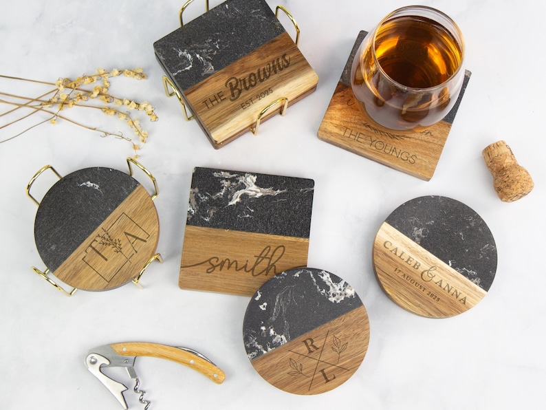 May include: A collection of square and circular coasters made of black marble and wood, featuring personalised engravings such as The Browns, The Youngs, Smith, Caleb & Anna, and initials. Includes a gold metal holder and a wine opener.