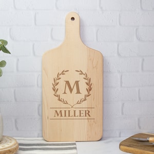 May include: A light-colored maple wood cutting board with a handle. The board features an engraved design of a laurel wreath surrounding the letter "M" and the name "MILLER" below. The board measures 17 inches by 8 inches.