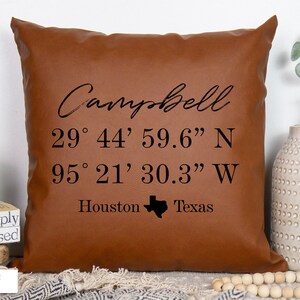 May include: Brown faux leather pillow with the text "Campbell" in cursive, followed by the coordinates "29° 44' 59.6" N 95° 21' 30.3" W", "Houston", and a small outline of the state of Texas.