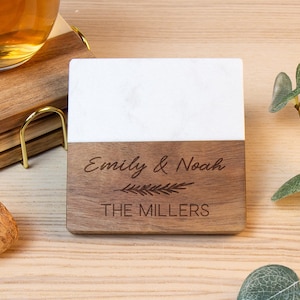 May include: A square coaster featuring a white marble top half and a dark wood bottom half. The wood section is engraved with the names Emily & Noah, a small leaf graphic, and the text THE MILLERS in a clean, sans-serif font.