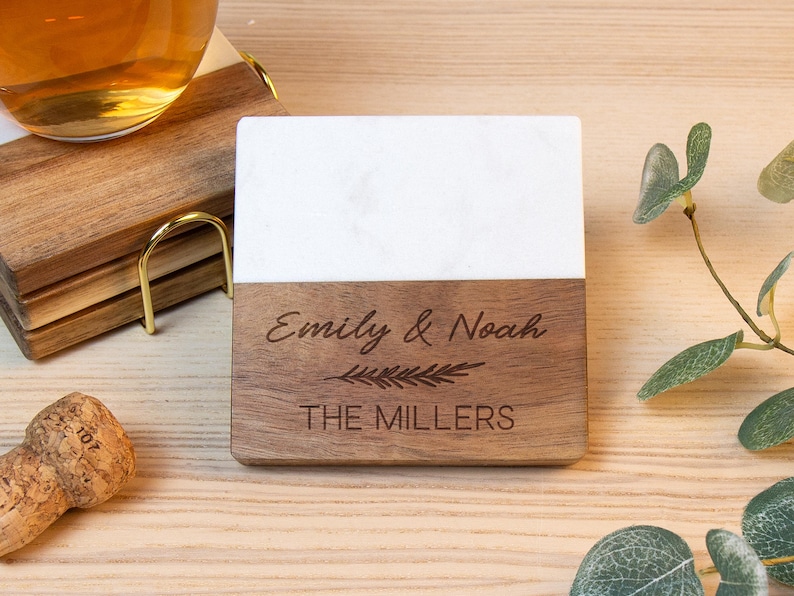 May include: A square coaster featuring a white marble top half and a dark wood bottom half. The wood section is engraved with the names Emily & Noah, a small leaf graphic, and the text THE MILLERS in a clean, sans-serif font.