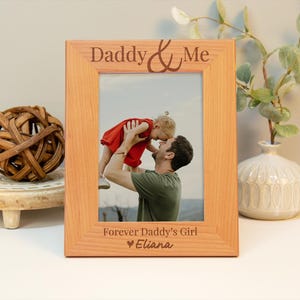 May include: A wooden picture frame with the text "Daddy & Me" and "Forever Daddy's Girl" with a heart and the name "Eliana". The frame has a photo of a father holding his daughter in the air.