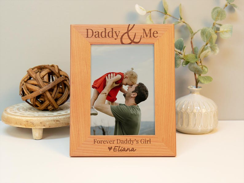 May include: A wooden picture frame with the text "Daddy & Me" and "Forever Daddy's Girl" with a heart and the name "Eliana". The frame has a photo of a father holding his daughter in the air.