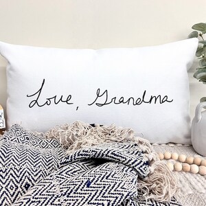 May include: White pillow with black cursive text that reads "Love, Grandma". The pillow is on a white surface with a blue and white blanket and wooden beads.