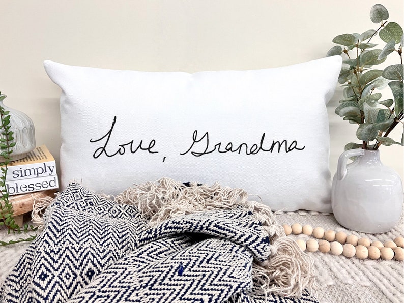 May include: White pillow with black cursive text that reads "Love, Grandma". The pillow is on a white surface with a blue and white blanket and wooden beads.