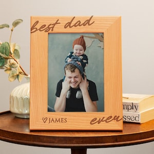 May include: A wooden picture frame with the text "best dad ever" and a heart with the name "JAMES" engraved on it. The frame has a photo of a father and child.