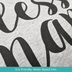 May include: Close-up of a gray sweatshirt with the word "bless" printed in black cursive lettering. The text is printed using eco-friendly, water-based inks.