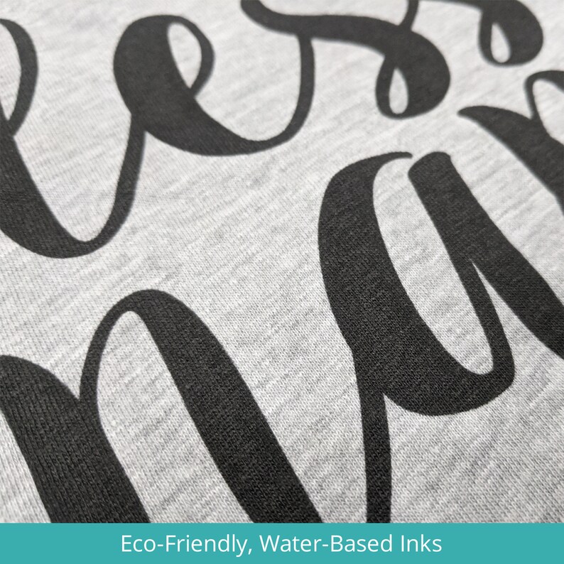 May include: Close-up of a gray sweatshirt with the word "bless" printed in black cursive lettering. The text is printed using eco-friendly, water-based inks.