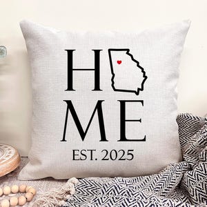 May include: White throw pillow with black text that reads "Home" with an outline of the state of Georgia and a red heart in the center. The text "ME" is below the state outline and the text "EST. 2025" is below that.