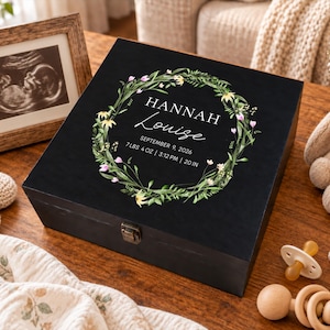 May include: A black wooden keepsake box featuring a floral wreath graphic surrounding the text HANNAH LOUISE, SEPTEMBER 9, 2026, 7 LBS 4 OZ | 3:12 PM | 20 IN. The box has a metal latch closure and a smooth, dark finish on the lid.