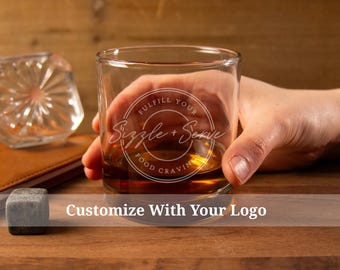 Peut inclure: Un verre à whisky en verre transparent avec un motif blanc qui dit "Fulfill Your Food Cravings Sizzle + Serve". Le verre est rempli d'un liquide brun et est tenu par une main.