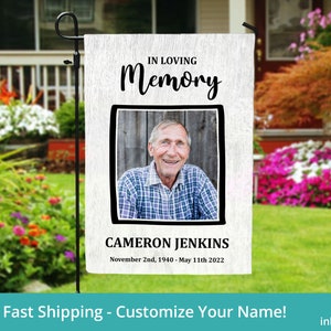 May include: A white garden flag with black text that reads "In Loving Memory" and a black framed photo of a man. The text below the photo reads "Cameron Jenkins, November 2nd, 1940 - May 11th 2022".