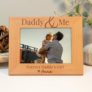 May include: A wooden photo frame with the text "Daddy & Me" and "Forever Daddy's Girl" with a heart and the name "Anna" engraved on it. The frame has a photo of a father holding his daughter in the air.