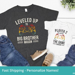 May include: Two t-shirts with a video game theme. The larger shirt is dark gray with the text "LEVELED UP TO BIG BROTHER MASON". The smaller shirt is white with the text "PLAYER 2 HAS ENTERED THE GAME CARSON".