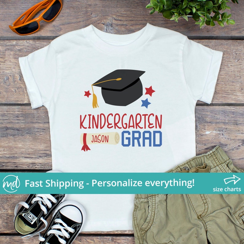 May include: White t-shirt with a black graduation cap, red and blue stars, and the words "Kindergarten Grad" in red and blue. The name "Jason" is printed below the words "Kindergarten" and "Grad". A rolled diploma is also printed on the shirt.