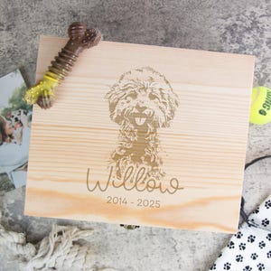 May include: A light wooden box with a laser-etched dog portrait and the name "Willow" and dates. A dog toy, tennis ball, leash, and photo are arranged around the box. The box is a pet-themed item.