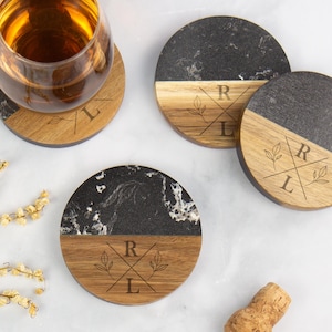 May include: Three circular coasters crafted from black marble and natural wood, each displaying a laser-engraved motif with the letters R and L divided by crossed lines and accompanied by small leaf drawings. One coaster holds a glass of amber liquid.