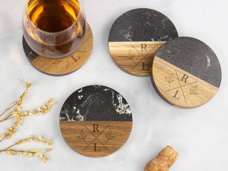 May include: Three circular coasters crafted from black marble and natural wood, each displaying a laser-engraved motif with the letters R and L divided by crossed lines and accompanied by small leaf drawings. One coaster holds a glass of amber liquid.