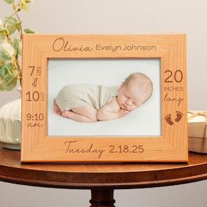 May include: A light wood photo frame with a baby photo inside. The frame has engraved text that reads "Olivia Evelyn Johnson", "7 lbs & 10 oz", "9:10 am", "20 inches long", and "Tuesday 2.18.25".