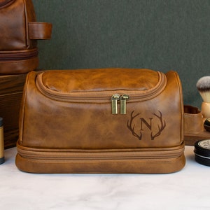 May include: A brown leather travel toiletry bag with a zipper closure and a monogrammed initial "N" with antlers. The bag has a top handle and a second zippered compartment.