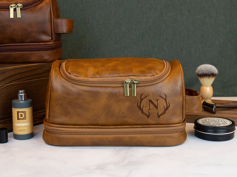 May include: A brown leather travel toiletry bag with a zipper closure and a monogrammed initial "N" with antlers. The bag has a top handle and a second zippered compartment.
