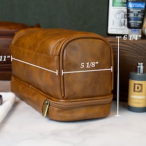 May include: Brown leather travel toiletry bag with two zippered compartments. The bag measures 11 inches wide, 6 1/4 inches tall, and 5 1/8 inches deep.