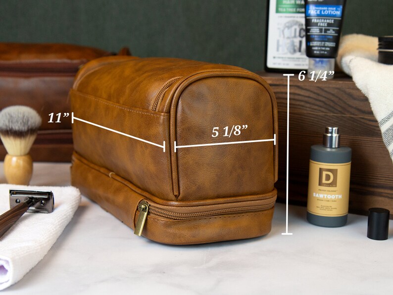 May include: Brown leather travel toiletry bag with two zippered compartments. The bag measures 11 inches wide, 6 1/4 inches tall, and 5 1/8 inches deep.