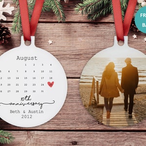 May include: White round ornament with a calendar for August with a red heart marking the 25th. The text "10th anniversary" and "Beth & Austin 2012" is below the calendar. The back of the ornament features a photo of a couple walking on a beach at sunset.